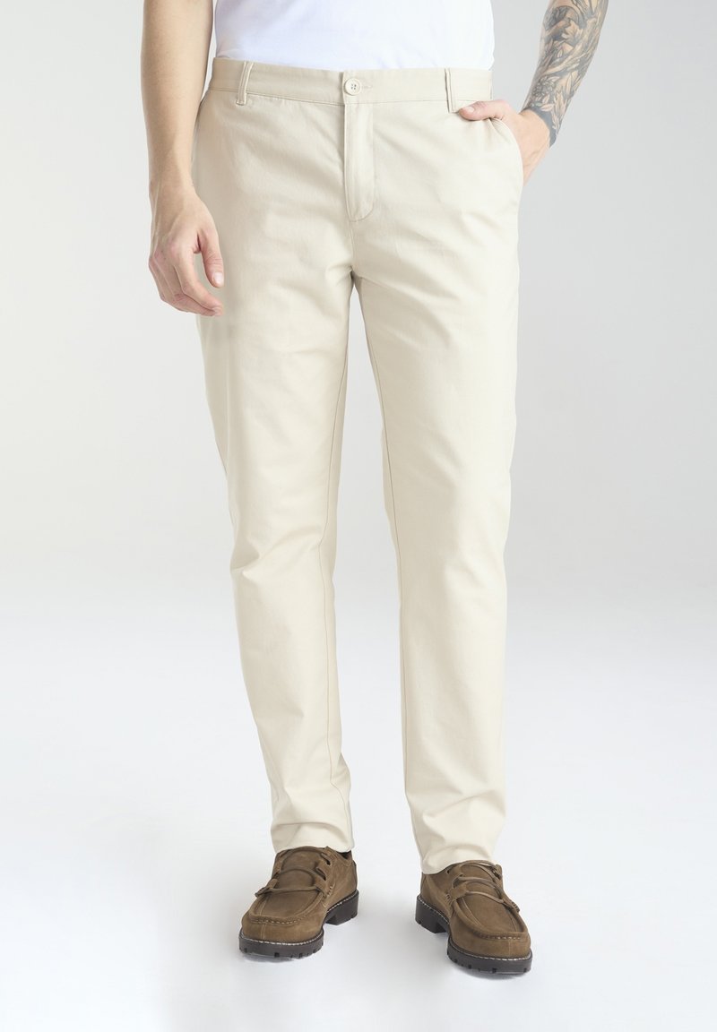 Beżowe, slim-fit spodnie z mieszanki bawełny, z pojedynczym zapięciem na guzik, kieszeniami z przodu i widocznymi detalami przeszyć, w połączeniu z brązowymi butami.