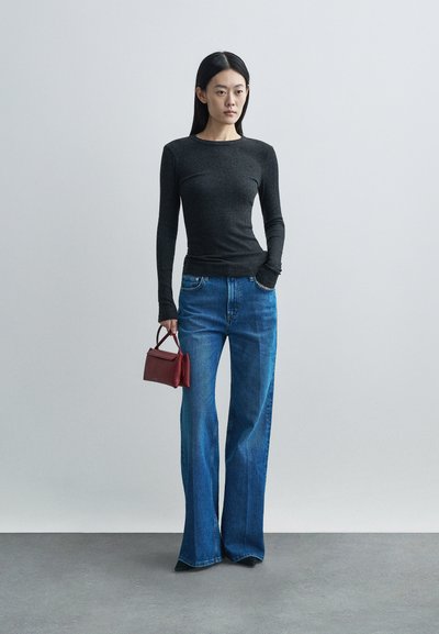 Γυναίκα που φοράει εφαρμοστό μακρυμάνικο μαύρο τοπ και παντελόνι με φαρδιά πόδια σε μπλε απόχρωση, κρατώντας μια μικρή κόκκινη τσάντα με το ένα χέρι στην τσέπη.