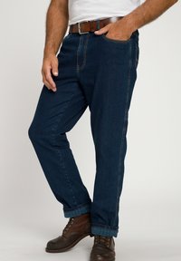 Jeans de mezclilla azul oscuro con corte recto, que cuentan con bolsillos frontales y un detalle de costura, combinados con botas de cuero marrón.