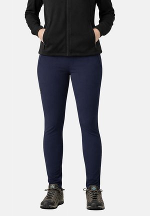 Personne portant un pantalon slim bleu marine, une veste zippée noire avec les mains dans les poches, et des chaussures de randonnée grises, debout devant un fond clair.