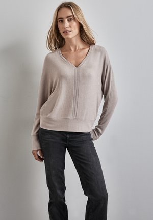 Hellbeige V-Ausschnitt-Pullover mit langen Ärmeln und gerippten Bündchen, kombiniert mit dunklen Jeans, vor einem neutralen Hintergrund.