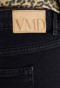 Black denim jeans with a visible leather tag labeled "VMID" stitched onto the waistband. Features include dark stitching and pocket outline.