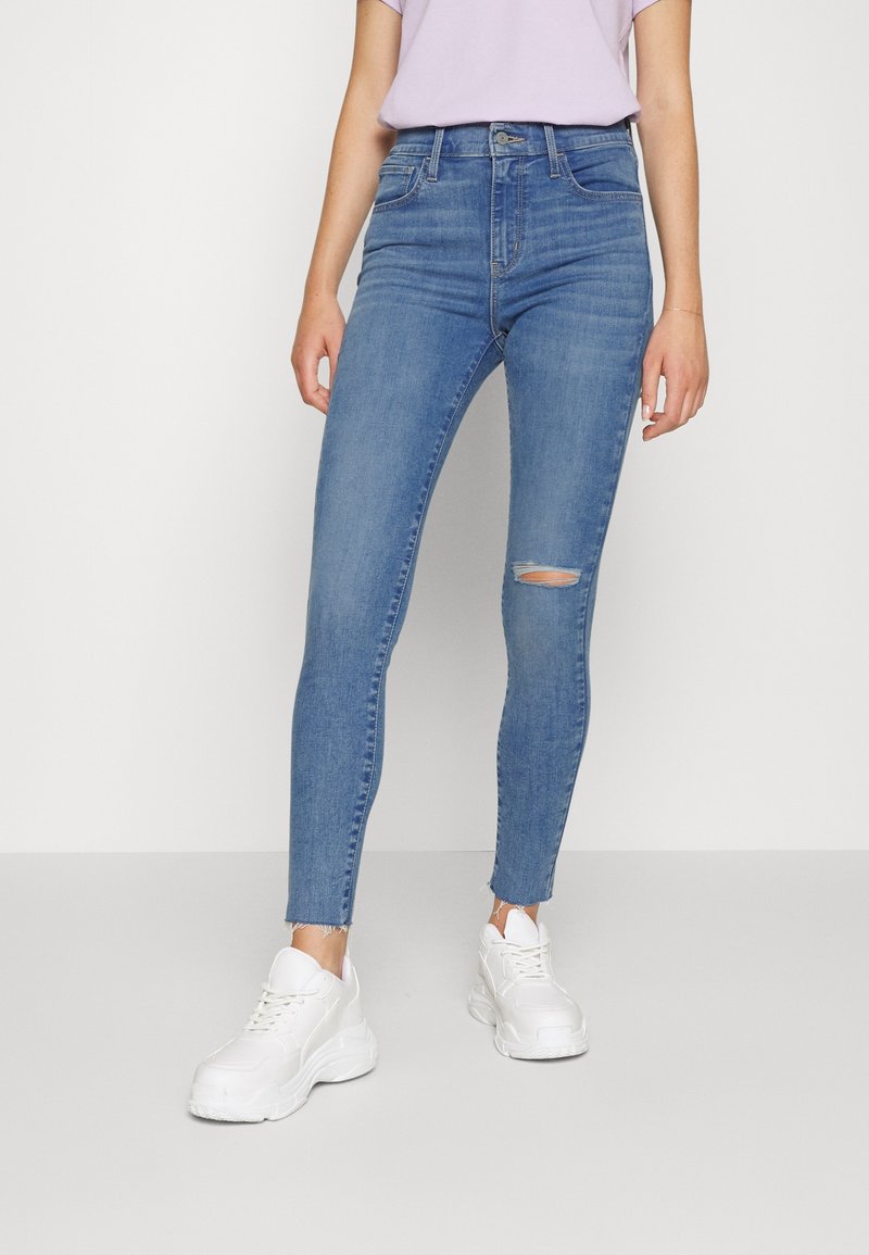 Jean slim en denim bleu clair avec une déchirure à l'avant et des chevilles effilochées, associé à des baskets blanches épaisses. Texture lisse avec un design classique à cinq poches.