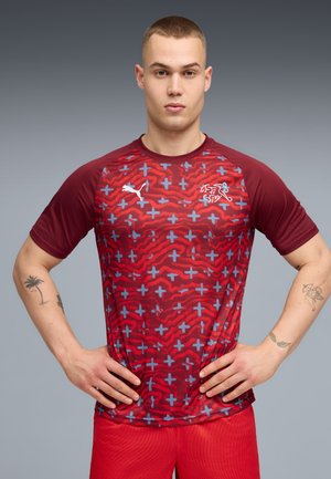 Fiatal férfi vörös sportruházatban, mintás inggel, magabiztosan állva, kezét csípőjén tartva, egyszínű háttér előtt.