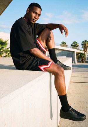 Młody mężczyzna ubrany w czarny oversize t-shirt, czarne szorty z czerwonymi i białymi paskami, czarne trampki, siedzący na betonowym murku na zewnątrz.