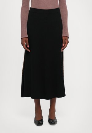 Frau in einem schwarzen gerippten Midikleid mit seitlichen Schlitzen, kombiniert mit einem mauvefarbenen Langarmoberteil und schwarzen Schuhen mit quadratischer Spitze.