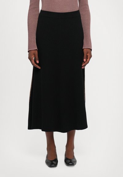 Donna che indossa una gonna midi a coste nera con aperture laterali, abbinata a un top a maniche lunghe color malva e scarpe nere con punta quadrata.