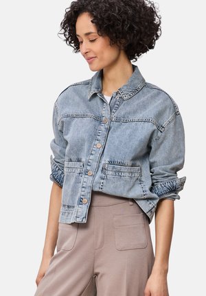 Kvinde med kort krøllet hår iført en lyseblå denimjakke med oprullede ærmer og beige højtaljede bukser, der kigger ned og smiler.