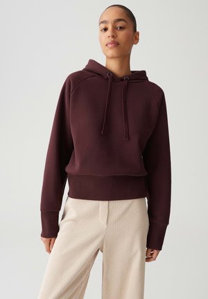Vrouw in een donker bordeauxrode hoodie met koordjes en crèmekleurige geribbelde broek, staand tegen een effen lichte achtergrond.
