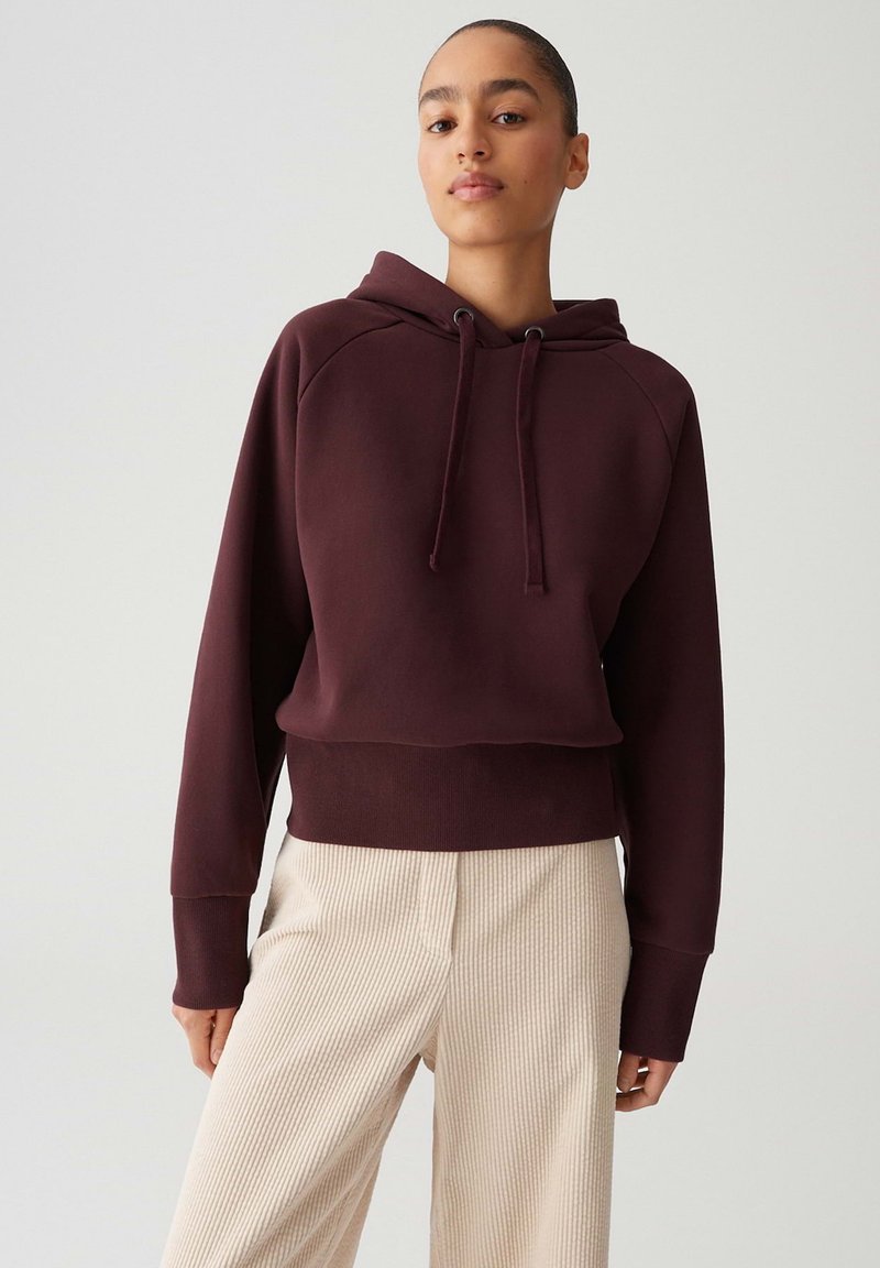 Donna che indossa un felpa con cappuccio color bordeaux scuro con cordini e pantaloni a coste di colore crema, che si trova di fronte a uno sfondo chiaro e semplice.