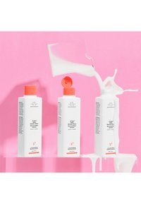 Drei weiße Flaschen mit orangefarbenen Kappen, beschriftet mit "E-Rase Milk Micellar Water", vor einem rosa Hintergrund, mit Milch, die aus einer Flasche spritzt.