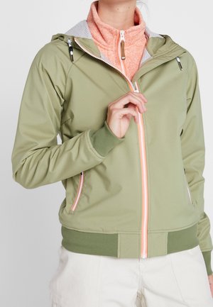 Persoon die een lichtgroene capuchonjas met roze rits dichtritst, draagt een perzikkleurige coltrui en witte broek.