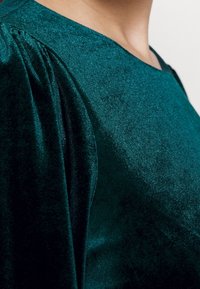 Smaragdgrön topp med en slät textur, rund halsringning och gathered puffärmar, tillverkad av ett mjukt, skimrande tyg.