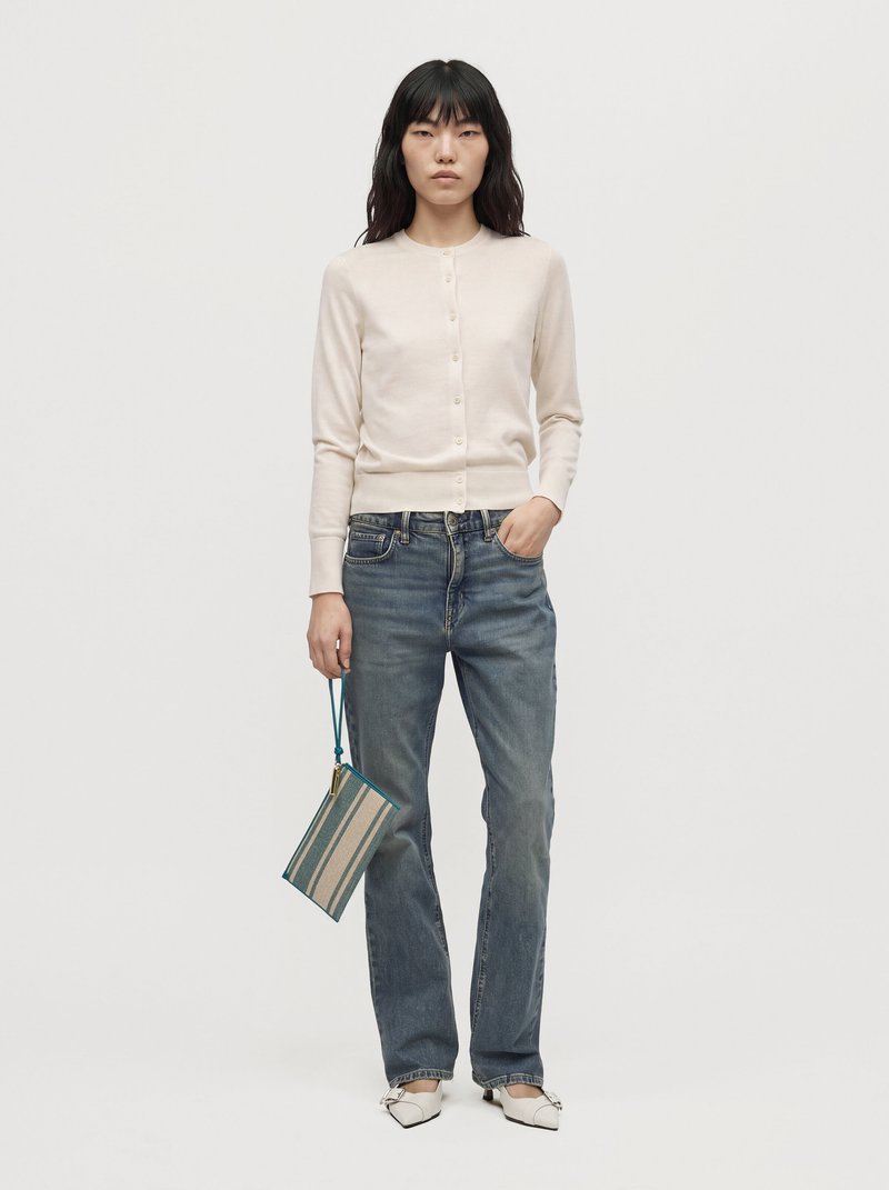Une femme porte un cardigan crème boutonné sur le devant, un jean bleu et des chaussures blanches. Elle tient une petite pochette rayée sur un fond de studio uni.