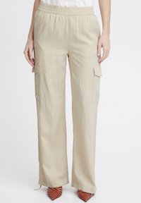 Pantalons cargo beiges avec une taille élastique, coupe ample, poches latérales et un tissu texturé. Portés avec des talons rayés orange et noir.