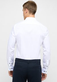 Mann in einem taillierten weißen Hemd und dunkelblauen Hosen, von hinten vor einem schlichten hellen Hintergrund abgebildet.