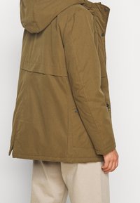 Olive green parka with a hood, featuring front pockets, a zippered side, and snap buttons. Made from textured fabric with a breathable design.