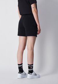 Pantaloni cargo neri con tasche laterali, vita elastica e texture liscia, abbinati a sneakers bianche e calzini a righe bianche e nere.