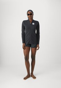 Black long-sleeve zip-up top with ribbed texture and a round logo patch. Paired with dark shorts, model stands barefoot against a plain background.