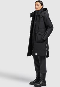 Woman standing wearing a long black puffer coat with pockets, hood, black pants, and black ankle boots on a plain light background.