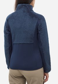 Femme portant une veste en molleton bleu marine avec un dos et des manches texturés doux, assortie à un pantalon beige, vue de dos.