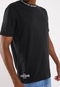 Camiseta de algodón negra con un cuello impreso en texto y un gráfico blanco "The ENERGY 1999" en el dobladillo. Mangas cortas y un corte relajado.