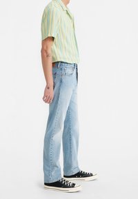Homme portant un jean bleu clair, des baskets basses noires et une chemise à manches courtes rayée pastel, debout de profil.