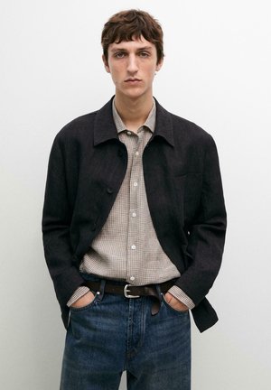 Junger Mann mit kurzen braunen Haaren, der eine schwarze Jacke und ein kariertes beiges Hemd trägt, das in blaue Jeans mit schwarzem Gürtel gesteckt ist, Hände in den Taschen.