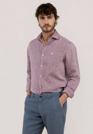 Hombre con pelo rizado que lleva una camisa de botones color púrpura claro y pantalones azules, con las manos en los bolsillos sobre un fondo liso.