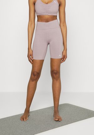 Kobieta stojąca boso na szarej macie do jogi, ubrana w jasnofioletowy marszczony sportowy biustonosz i wysokie krótkie spodenki rowerowe z logo Puma.