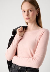 Mlada žena s ravnom svijetlosmešavom kosom, obučena u ružičastu majicu dugih rukava i crne traperice, drži crnu jaknu preko ramena.