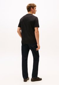 Mladenič s kodrastimi lasmi stoji obrnjeno stran, oblečen v črno majico s kratkimi rokavi, temne kavbojke in rjave usnjene loaferje.