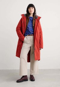 Cappotto puffer lungo rosso con cappuccio, foderato in grigio e con chiusure a scatto, indossato sopra una camicia in corduroy marrone e una maglietta a righe blu, abbinato a pantaloni in corduroy beige e stivali bordeaux.