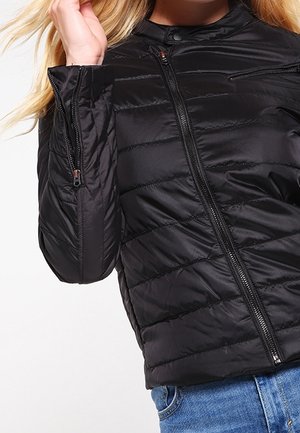 Femme portant une veste matelassée noire avec des fermetures éclair devant et aux manches, associée à un jean bleu, sur un fond uni.