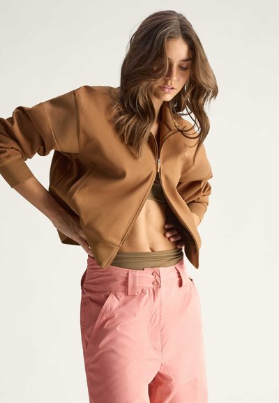 Braune Zip-Jacke mit gerippten Bündchen, kombiniert mit hoch taillierten rosa Hosen und einem olivgrünen Oberteil mit glatter Textur und lässigem Schnitt.