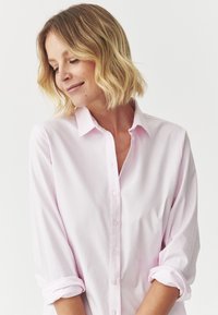 Chemise boutonnée rose clair avec col, manches longues retroussées. Tissu lisse avec un léger éclat, comportant une seule rangée de boutons sur le devant.