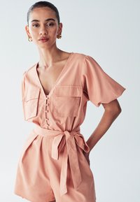 Peegli värvi kombekas lühikeste varrukatega, V-kaelusega, nööpide esiava, kahe rinna tasku ja seotud taljevööga. Sile kanga tekstuur.