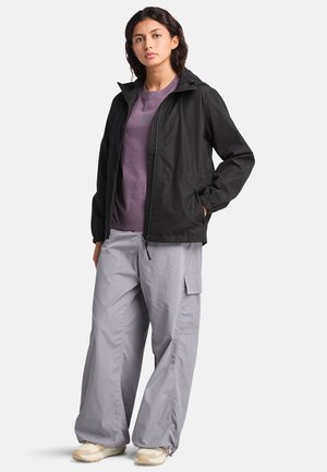 Chaqueta ligera negra con capucha, llevada sobre una camisa morada. Combinada con pantalones cargo grises holgados y zapatillas beige.