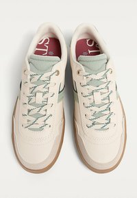 Beige leather sneakers with green and navy accents, textured mint green collar, and patterned laces on a light brown rubber sole.