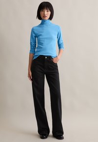 Hellblaues Rollkragenoberteil mit langen Ärmeln, kombiniert mit hoch taillierten schwarzen weiten Jeans. Das Model steht mit einer Hand in der Tasche.
