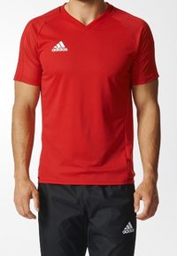 Hombre vestido con una camiseta deportiva de manga corta roja de Adidas y pantalones atléticos negros de Adidas, contra un fondo liso.
