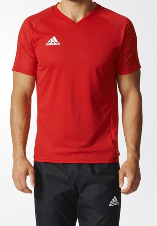 Hombre vestido con una camiseta deportiva de manga corta roja de Adidas y pantalones atléticos negros de Adidas, contra un fondo liso.