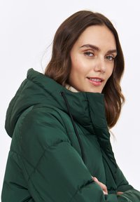 Zielona kurtka puchowa z kapturem, o pikowanej strukturze i czarnym sznurkiem. Widoczny beżowy golf pod spodem.