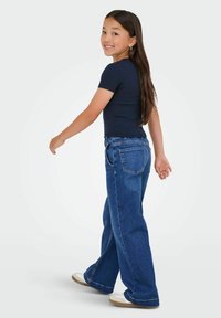 Fille aux longs cheveux noirs portant un t-shirt bleu marine, un jean large bleu et des baskets blanches, marchant et souriant par-dessus son épaule.