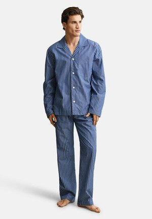 Man staat op blote voeten en draagt een pyjamajasje met lange mouwen in blauw-witte verticale strepen, gecombineerd met bijpassende broek.