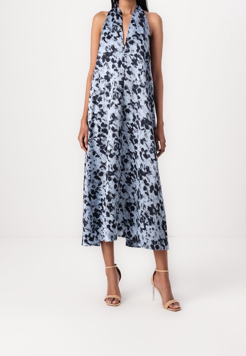 Femme portant une robe midi sans manches, à motifs bleus et noirs, avec un décolleté en V profond et des sandales à talons hauts couleur nude, sur un fond blanc.