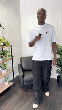 Un tricou alb cu mânecă scurtă și grafică roșie, pantaloni scurți negri și pantofi albi, stând într-o cameră cu plante și mobilă.