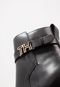 Musta nahkasaapas nilkkamittaisena kiiltävällä pinnalla, jossa on kultasävytteinen "TH" logo hihnassa ja ompelut yksityiskohdat sivulla.