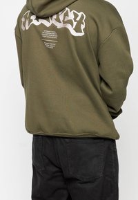 Felpa verde oliva con un grande graphic design testurizzato bianco sul retro. Presenta un cappuccio con cordino e una vestibilità rilassata. Pantaloni neri visibili.