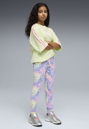 Nastolatka stoi z założonymi ramionami, ubrana w oversize'ową pastelowożółtą koszulę, wielokolorowe legginsy w różowo-niebiesko-żółte pasy oraz srebrne trampki na szarym tle.